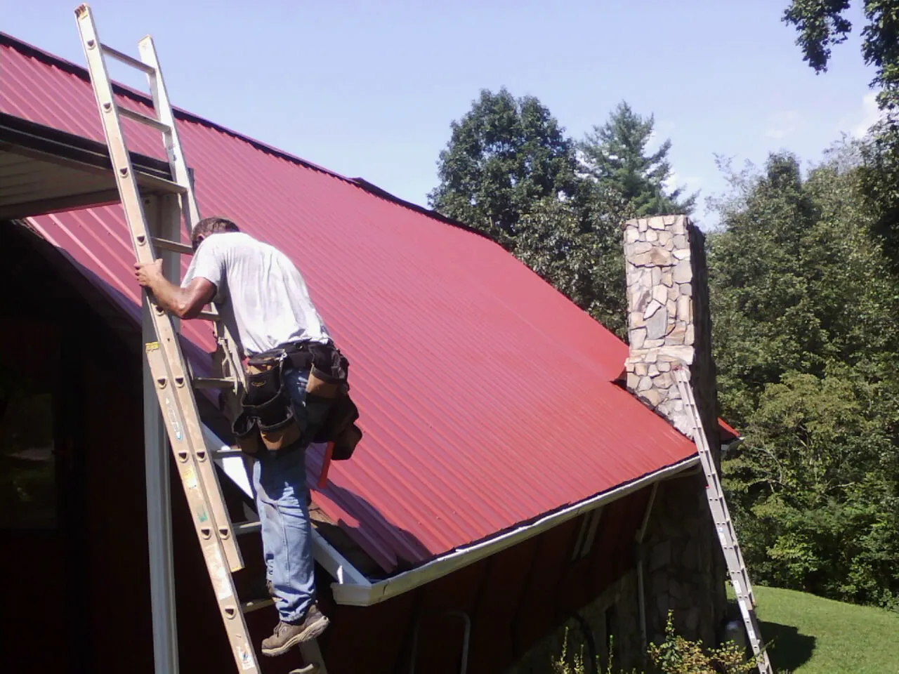 Roofer on a ladder beside a red metal roof and stone chimney — Soffit & Fascia Repair by Roofing Brothers in Bellevue